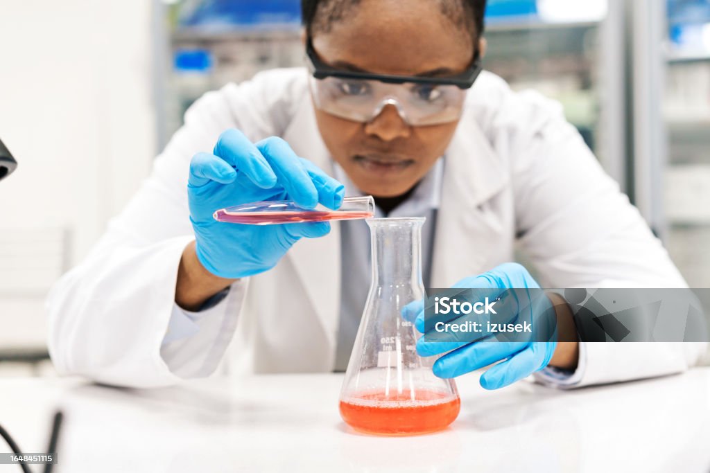 Focused female scientist pouring chemical in beaker while working at laboratory Scientist Stock Photo