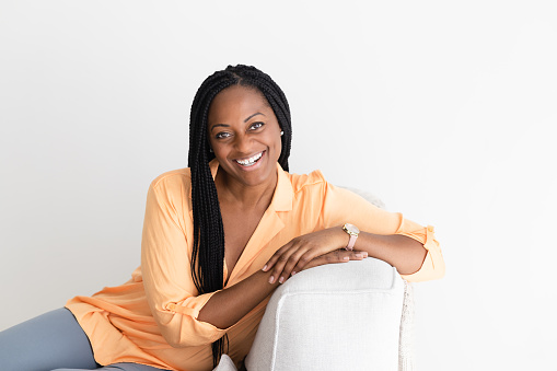 Black woman in her 40s, with stylish braids, sits on a comfortable sofa, her gaze focused and confident as she looks into the camera.