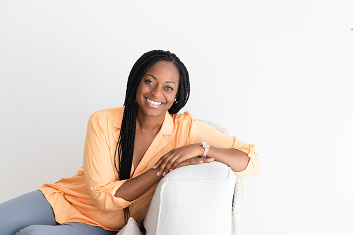 Black woman in her 40s, with stylish braids, sits on a comfortable sofa, her gaze focused and confident as she looks into the camera.