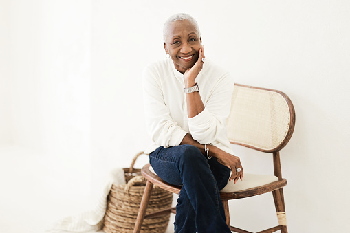 poised Black woman, elegantly dressed in a white shirt, sits on a chair, her gaze fixed on the camera, radiating confidence and grace.
