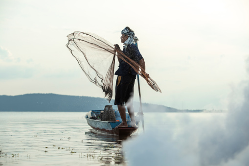 Middle aged Asian fisherman while throwing fishing net from boat on the lake, Thailand countryside.