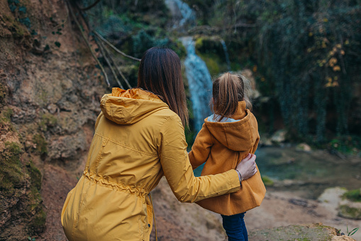 Mother and daughter standing in nature and watching the waterfall in autumn, dressed warmly.
