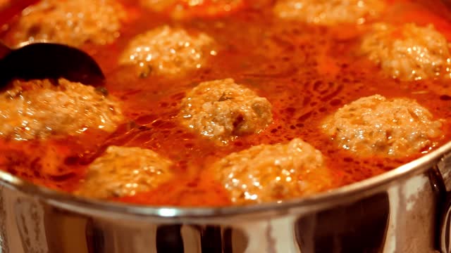 Cooking meat balls in tomato sauce. Close-up, selective focus. A hand stirring homemade meatballs boiling in a pan with tomato sauce. Delicious homemade beef and pork meat balls with rice being boiled in cooking pan with tomato sauce