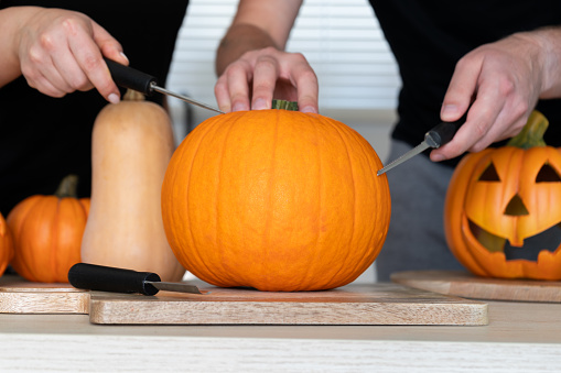 Couple preparing to carve Halloween pumpkins, spooky Jack-o'-lantern cut out. Man and woman standing behind uncarved pumpkin on a table. They holding carving tools, knives, carvers, saw knife blades.
