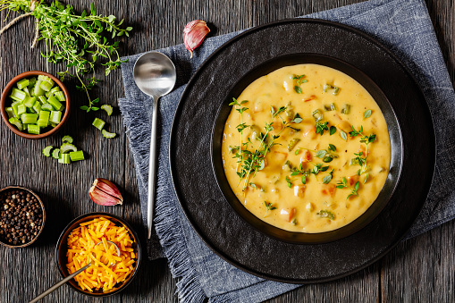 Beer cheese soup with vegetables and thyme in black bowl on dark wooden table with ingredients, spoon and tea towel, horizontal view from above, flat lay