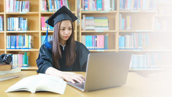 An asian girl student in university graduate gown with books, laptop studies happily in library.