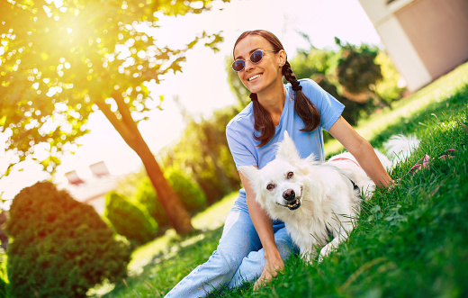 Happy young pretty brunette girl hugging her cute white fluffy dog on the outdoor in the park on lawn. Adoption, rescued, shelter, companion, pet.