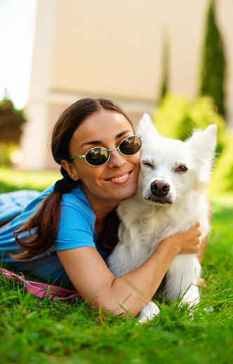 Happy young pretty brunette girl hugging her cute white fluffy dog on the outdoor in the park on lawn. Adoption, rescued, shelter, companion, pet.