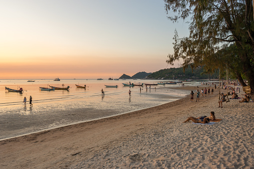 Koh Tao Island, Thailand - March 22, 2023: People and tourists walks on shore and rest on sandy Sairee beach and watching the sunset and look on sea during the low tide in the evening