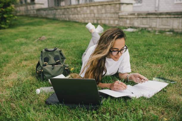 Young woman student studying using her books, laptop and notebook, sitting on grass.