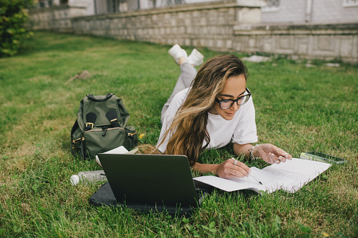 Young woman student studying using her books, laptop and notebook, sitting on grass. Back to school.