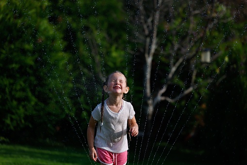 A smiling young girl enjoying the summertime by running and playing around a garden sprinkler