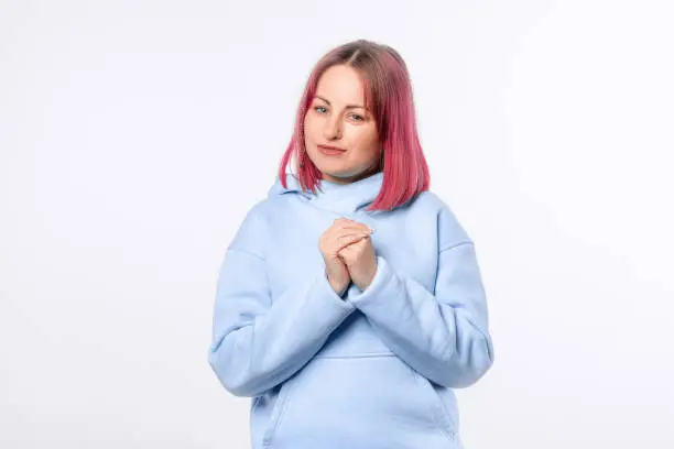 Pretty woman say thank you, express gratitude, looking cute and flattered, holding hands on heart. Lovely female model with pink hair in a blue hoodie standing over white background