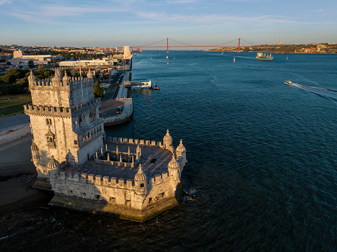 Aerial view of Belem Tower famous tourist landmark of Lisboa and tourism attraction on the bank of the Tagus River (Tejo) with tourist boat and cargo container ship at sunset. Lisbon, Portugal