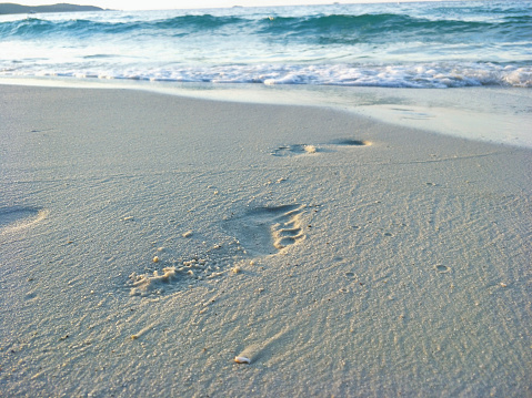 Footprints lead across the sand of an idyllic beach, back lit by the sun at dawn or dusk.