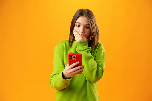 Surprised teen girl using mobile phone against yellow background close up