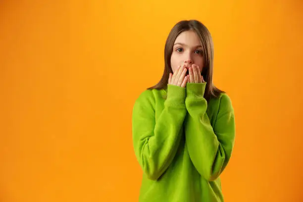 Teen girl over yellow background in studio shocked covering mouth with hands