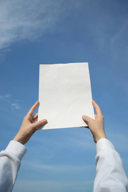 istock Hands holding up a blank sheet of paper outdoors, on a blue sky background. 1648770910