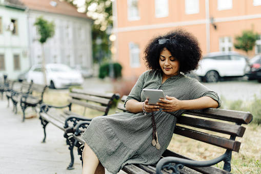 Young afro woman sitting on park bench and using digital tablet