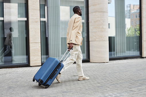 Rear view of young African American businessman in formalwear pulling suitcase with baggage while hurrying to airport or train station