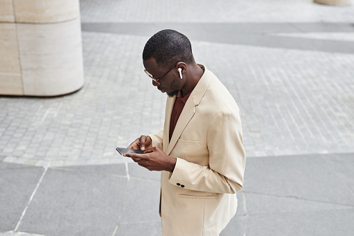 High angle of young black businessman in earphones and pastel color suit texting in mobile phone or scrolling through playlist in the street