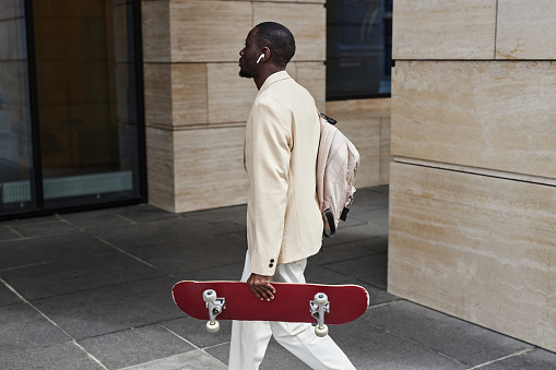 Side view of young African American solopreneur or office worker with backpack on shoulder and skateboard in hand walking to work
