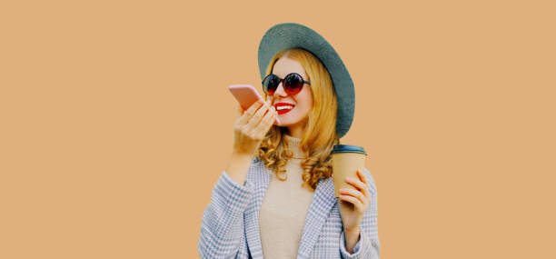 istock Beautiful stylish young woman with coffee cup holding smartphone using voice command recorder, assistant or takes calling wearing coat, round hat on brown background 1648844028