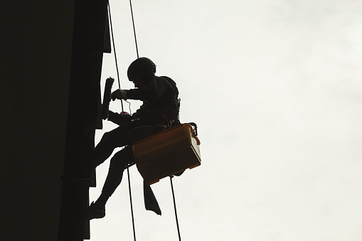 Silhouette of industrial mountaineer worker in uniform and helmet hanging wall, washing fasade, working on building. Access laborer high-rise works. Industry urban works concept. Copy ad text space