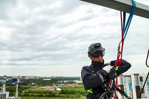 Male industrial mountaineer worker in uniform and helmet hanging over horizon, working on building. Access laborer high-rise rope dangerous works. Industry urban works concept. Copy ad text space