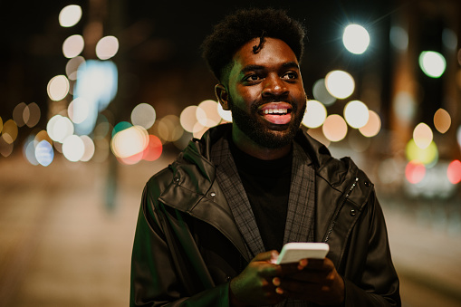 Portrait of a happy urban African American man standing on the street downtown at night and using his phone. A happy man is standing on the street and using cutting-edge technologies for communication