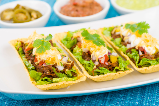 Shredded beef taco trays topped with salsa, sour cream and grated cheese. Jalapenos, refried beans and guacamole on background.