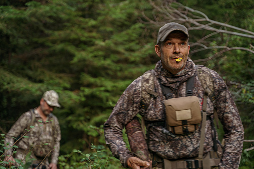 Middle aged Caucasian hunter hikes a forested mountain trail, looking for elk as a fellow hunter hikes behind him in the Pacific Northwest.