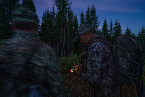 Two middle aged men wearing camouflage clothing stand in the forest at evening time, looking down at a tracking device while crossbow hunting in Washington State.