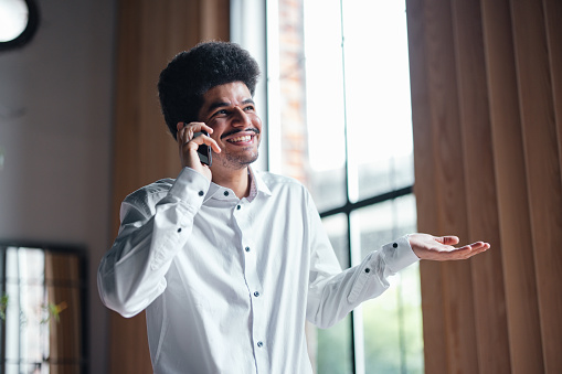A smiling Latin-American entrepreneur chatting on his smartphone while  standing in the office.