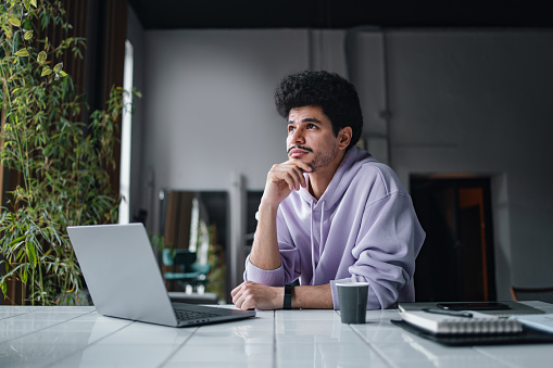 A serious Latin-American entrepreneur looking away and thinking while sitting at his office desk, in front of his laptop.
