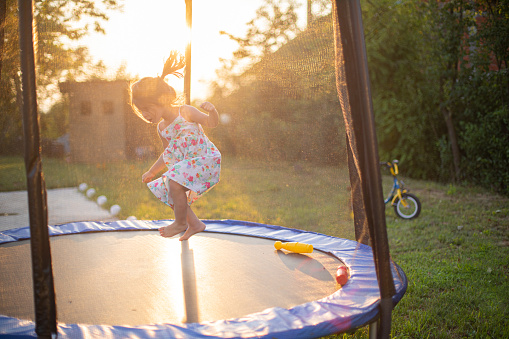 Three children jump on an outdoor trampoline with a safety net in the backyard. The shot is focused on a nine-year old boy in the front.