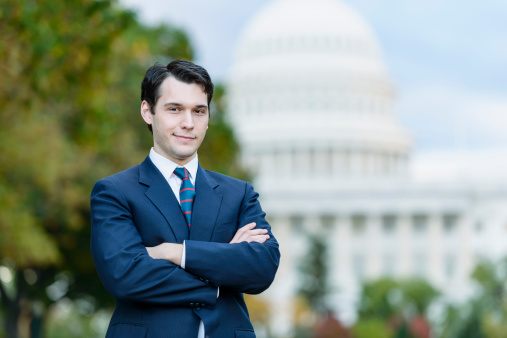 A young politician, staffer, or lobbyist holds with his arms folded, with a confident smile. The United States Capitol building is in the background.