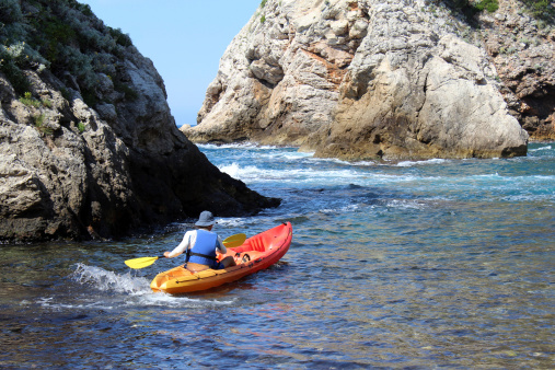 Colourful photo of a man kayaking in a orange and yellow kayak in Dubrovnik, Croatia on the Adriatic Sea.  Kayaker is about to kayak through a narrow water passage way of rapids, between two rock cliffs.  Photos was taken right outside the Old Walled City in a small rocky bay.