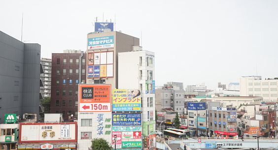 Kita City, Japan - June 10, 2023: Buildings and commercial signs line the downtown district outside Oji Station. Pedestrians walk on the elevated Oji Ekimae Pedestrian's Bridge over traffic on Route 122 and Route 501.  Spring afternoon with hazy skies in the Tokyo Metropolis.