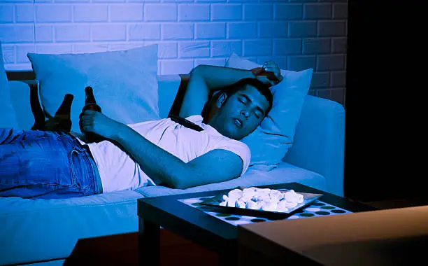 A man sleeps on the couch, with a TV remote control balancing on his protruding stomach.