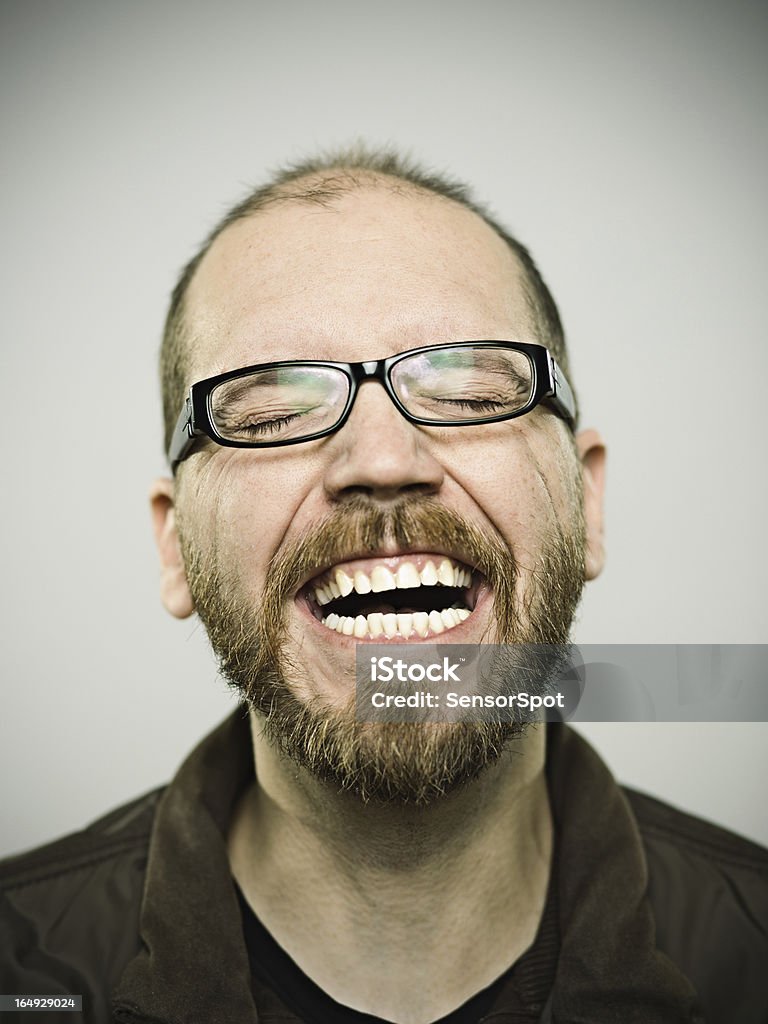 Man having a good laugh over gray background A young Caucasian man tilts his head back and smiles widely.  He is wearing glasses and has his eyes closed.  He has a moustache and beard. Mug Shot Stock Photo