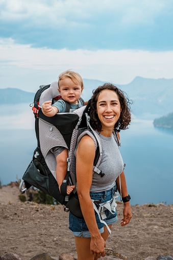 A beautiful and fit Eurasian woman smiles while hiking around Crater Lake with her one year old son who is riding in a backpack baby carrier.