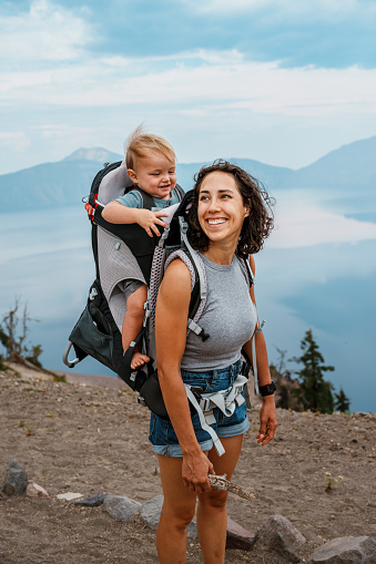 A beautiful and fit Eurasian woman smiles while hiking around Crater Lake with her one year old son who is riding in a backpack baby carrier.