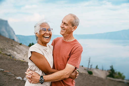 A vibrant senior woman of Hawaiian and Chinese descent laughs and affectionately embraces her Caucasian husband while out on a mountain hike overlooking a lake.