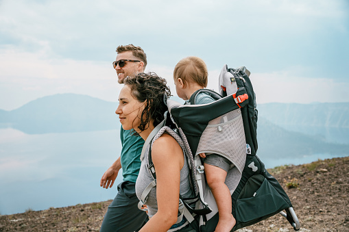 Profile view of an Eurasian woman carrying her one year old baby son in a backpack baby carrier while on a hike with her Caucasian husband overlooking Crater Lake in Oregon on a sunny and warm summer day.