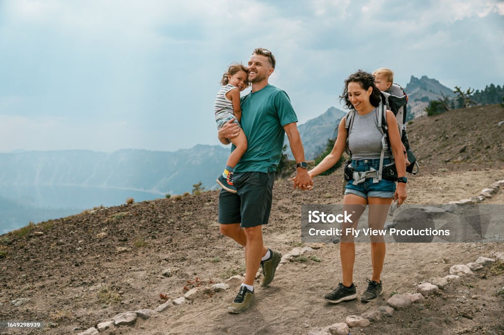 Family hiking in the mountains A fit young dad carries his three year old daughter and holds hand with his multiracial wife who is carrying their one year old son in a backpack baby carrier during a hike overlooking Crater Lake in Oregon. Family Stock Photo Family hiking in the mountains A fit young dad carries his three year old daughter and holds hand with his multiracial wife who is carrying their one year old son in a backpack baby carrier during a hike overlooking Crater Lake in Oregon. Family Stock Photo