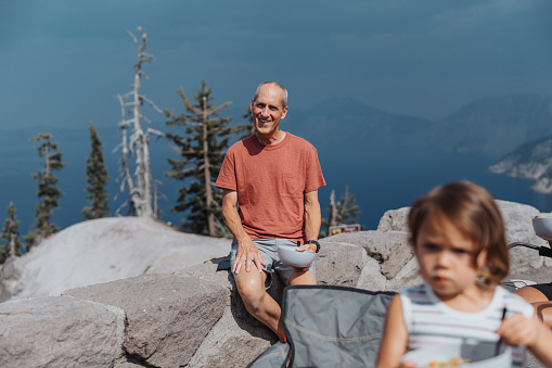 Portrait of an active and healthy senior man eating lunch at a scenic viewpoint overlooking Crater Lake in Oregon on a sunny and warm summer day. The man is on vacation with his kids and grandkids.