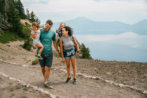 An active and fit multiracial couple with a toddler daughter and one year old baby son cheerfully talk while on a hike overlooking Crater Lake during a summer camping road trip through Oregon.