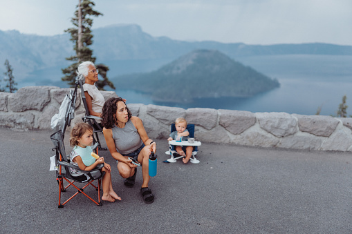 A multiracial multi-generation family sits in camping chairs and eats a picnic lunch at a scenic viewpoint overlooking Crater Lake in Oregon during a summer camping road trip.