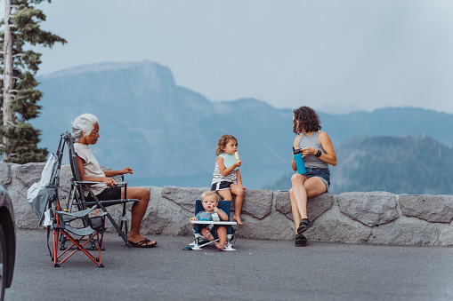 An Eurasian woman sits on a rock wall and enjoys the view of Crater Lake while on a road trip with her vibrant senior adult mother who is of Pacific Islander descent and her young children. The woman's toddler daughter is eating a popsicle and her one year old son is sitting in a booster seat eating a snack.